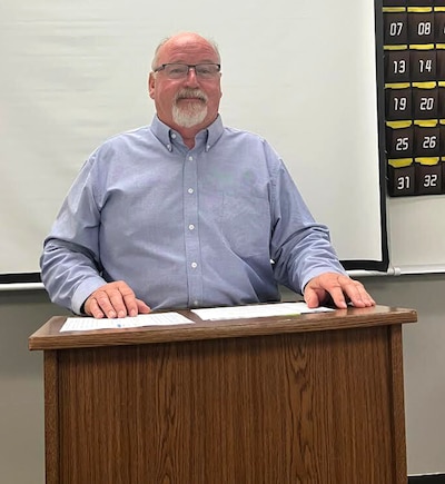 A man in a blue shirt and glasses stands behind a podium