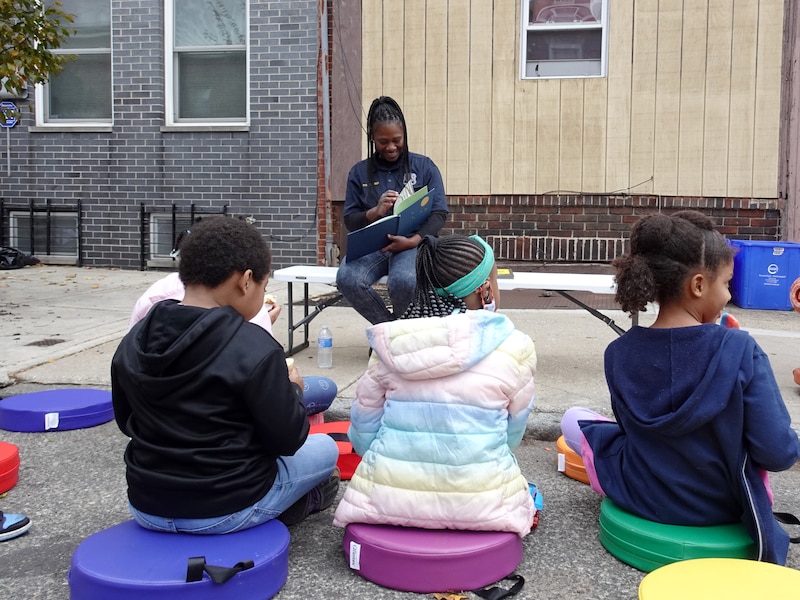 A person with long dark hair sits on a sidewalk reading to three children who are sitting on colorful cushions.