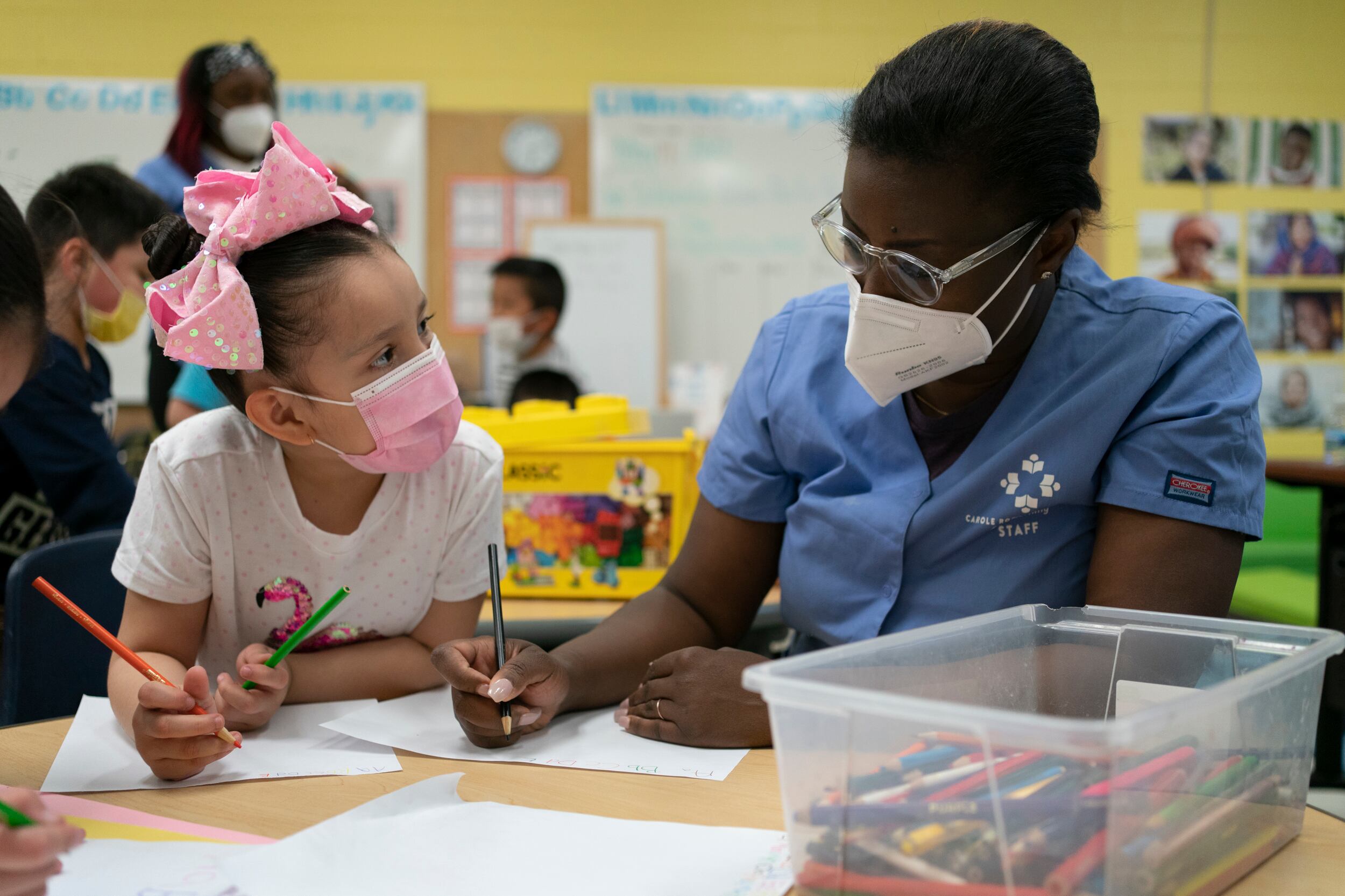 An early childhood student support worker in a blue shirt, wearing a filtered mask assists a girl wearing a pink bow and pink protective mask.