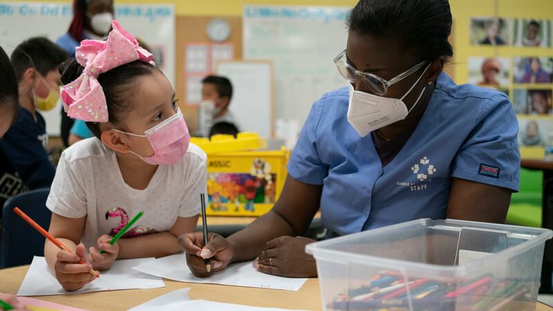 An early childhood student support worker in a blue shirt, wearing a filtered mask assists a girl wearing a pink bow and pink protective mask.