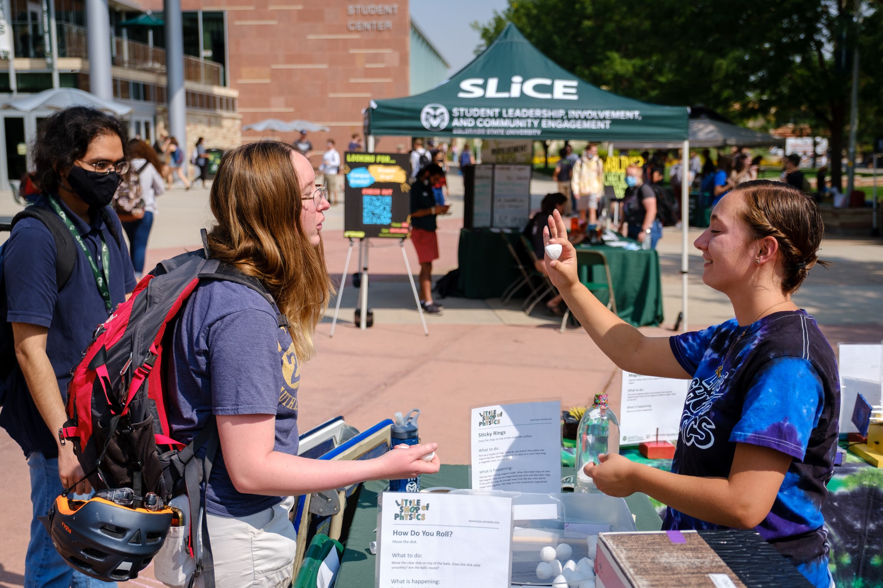 Three young adults stand outside a university building with a green tent in the background.