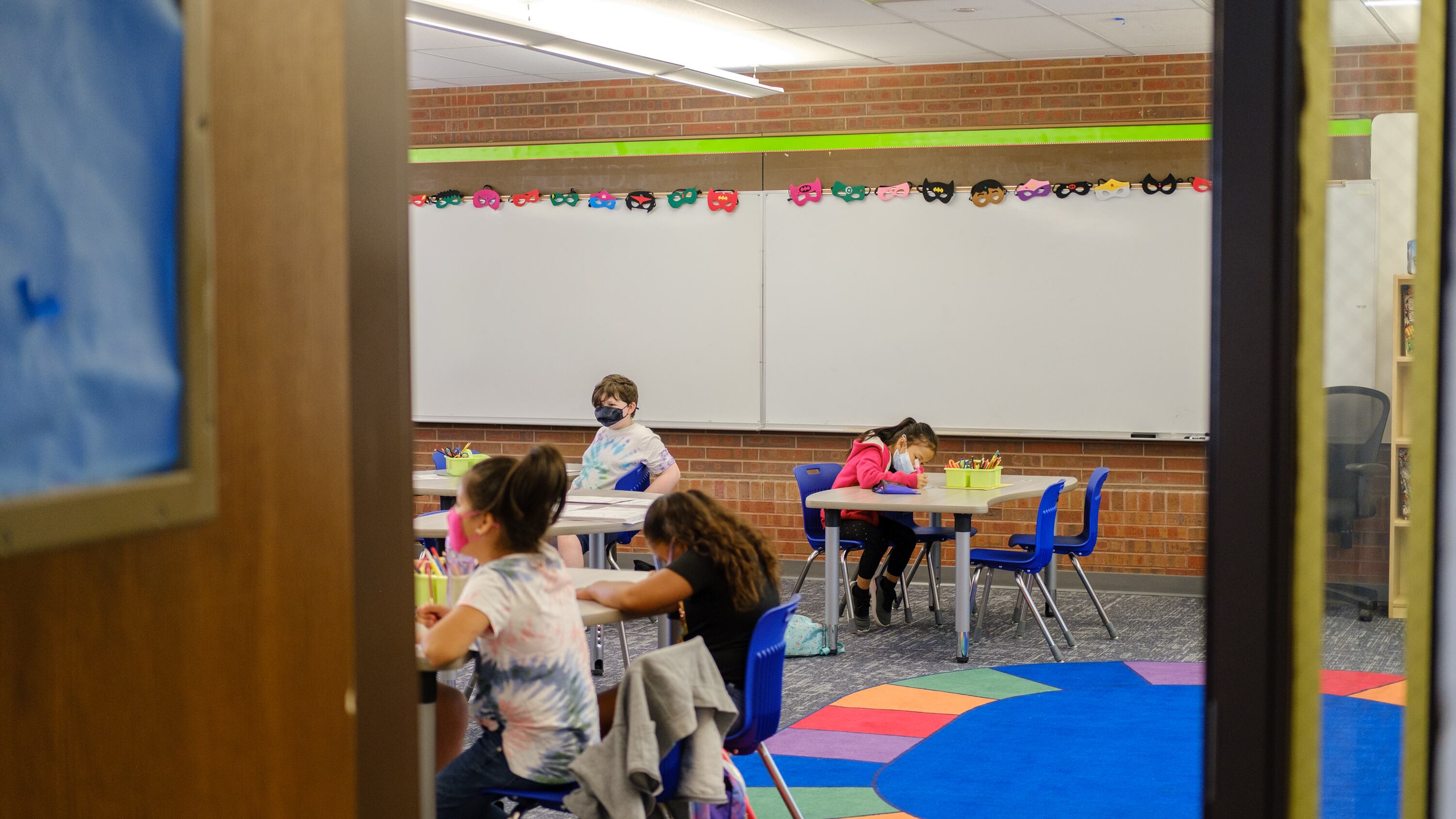 Students sit at tables in a classroom, with a colorful circle-shaped rug dominating the photo.