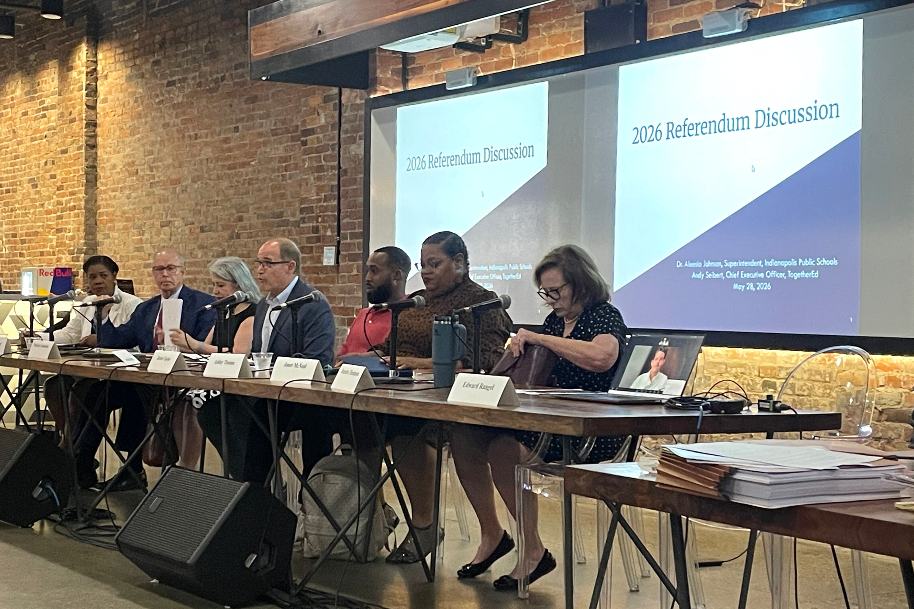A photograph of seven adults sitting at a long table with two large projector screens that read "2026 Referendum Discussion."