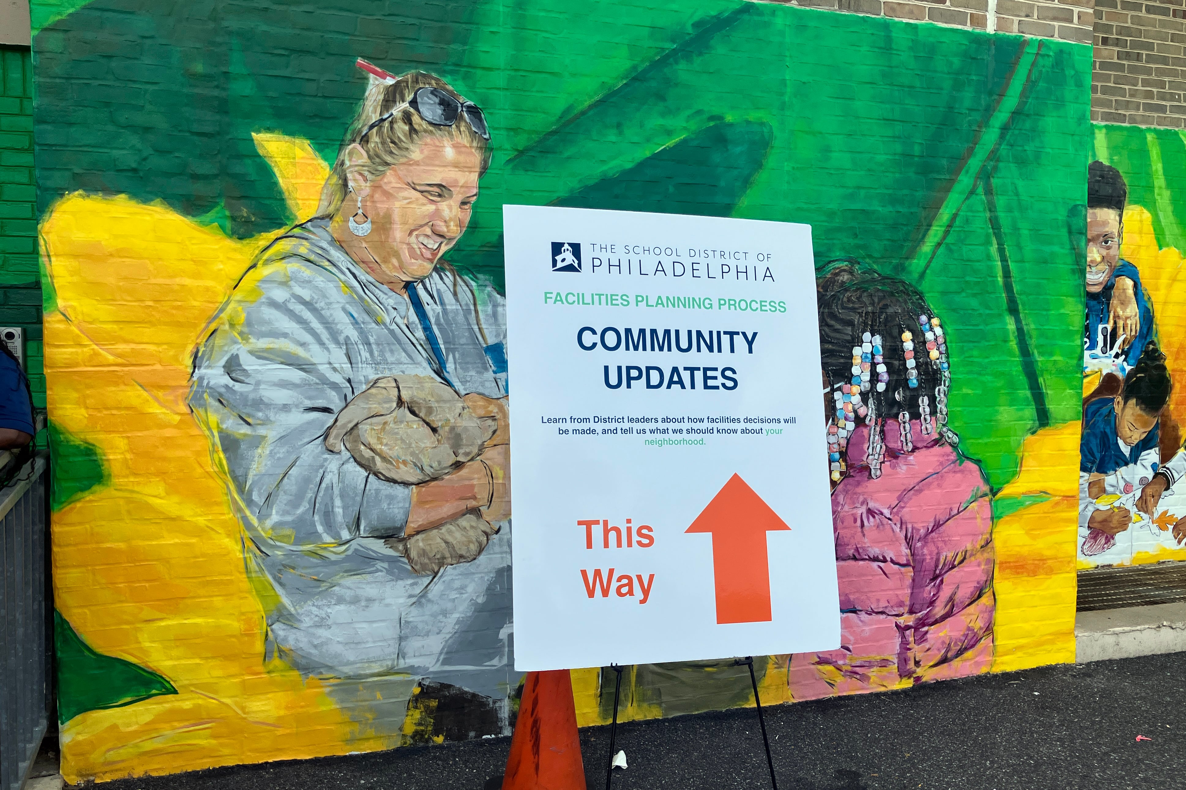 A photograph of a poster board standing in front of a mural outside of a school building.