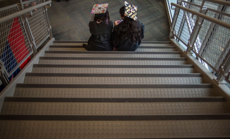 Two high school graduates in caps and gowns sit on a staircase.
