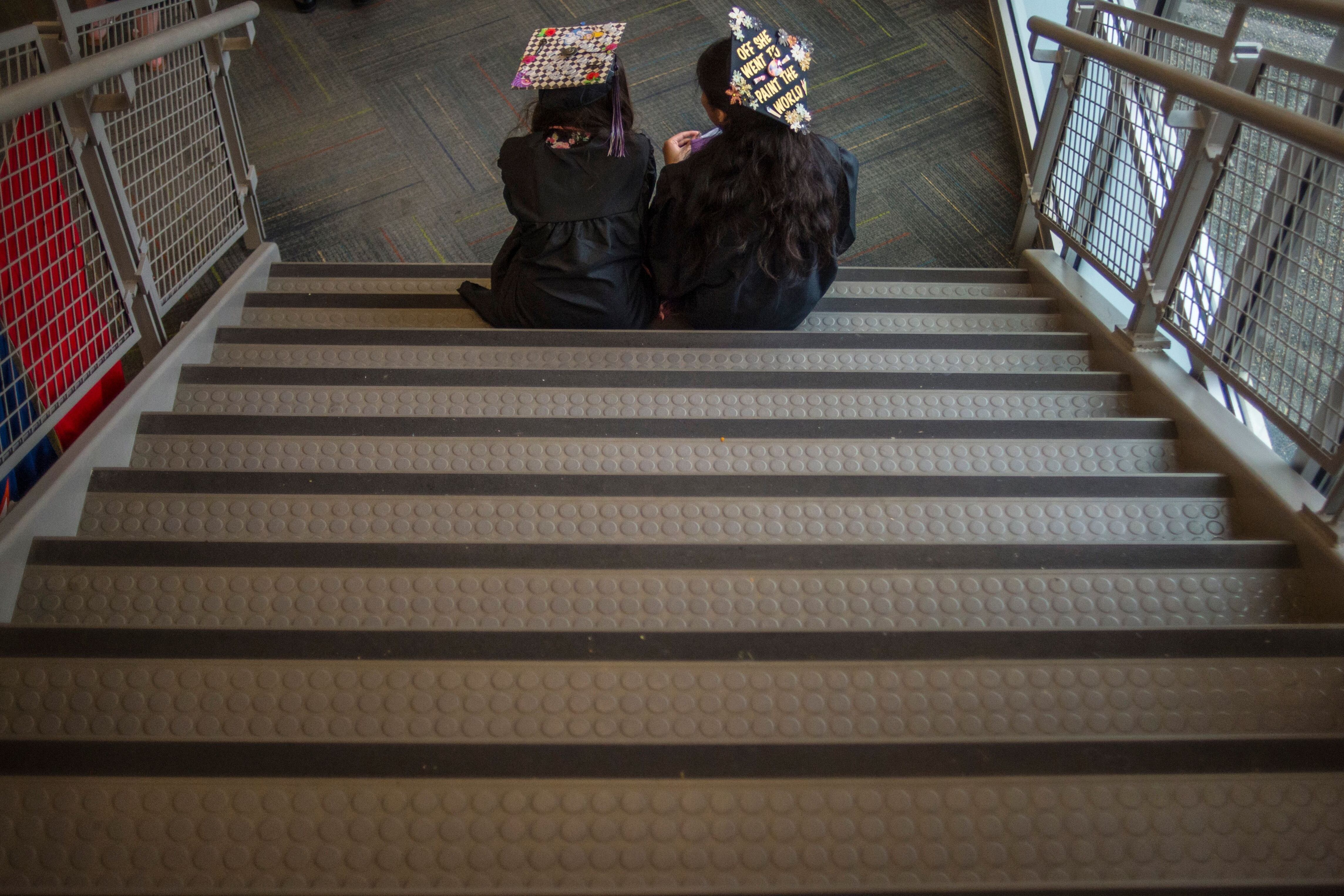 Two high school graduates in caps and gowns sit on a staircase.