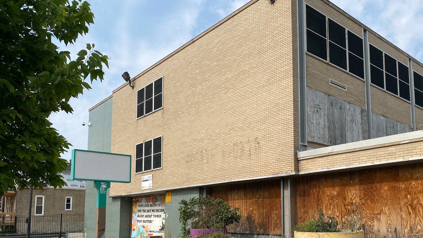 A tan brick and green tile school building with a blank sign sits empty and boarded up. Four orange, purple, and yellow planters line the sidewalk in front of the building.