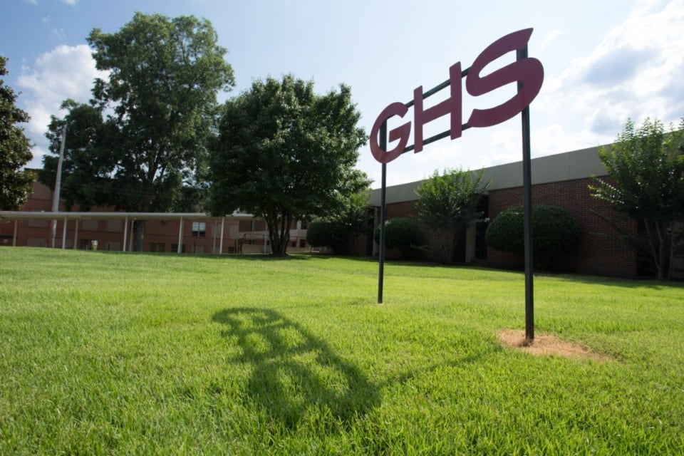 A sign that says “GHS” sits in front of a school building surrounded by a green campus.