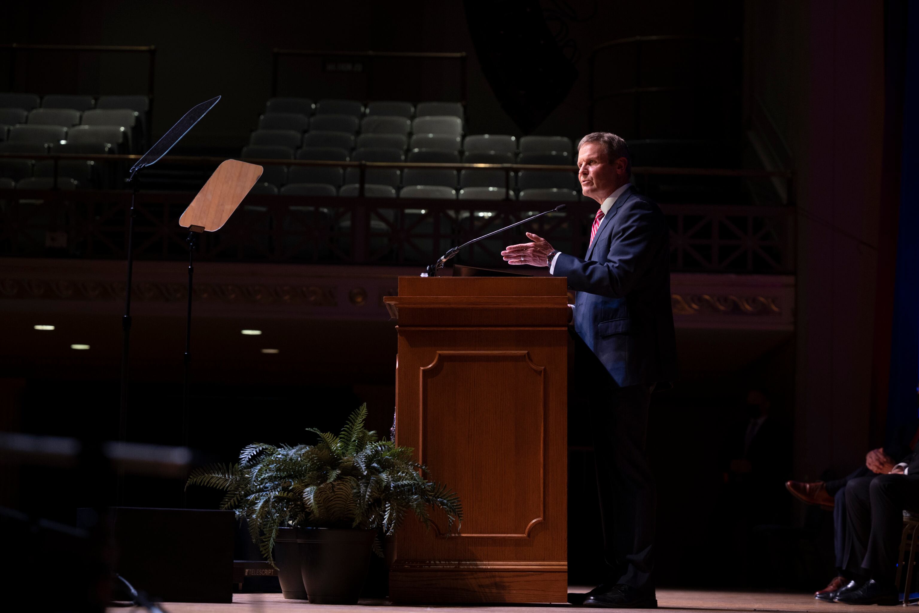 Tennessee Gov. Bill Lee wears a blue suit and stands on stage behind a wooden podium in a large assembly hall with empty seats.