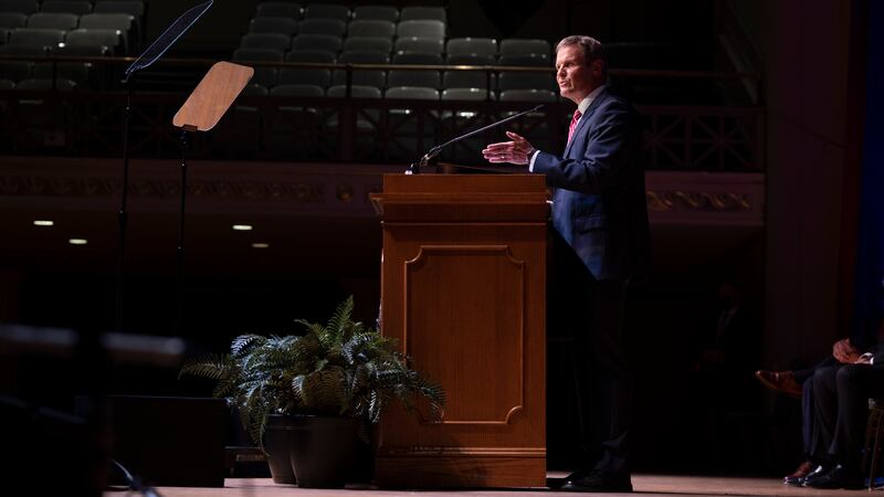 Tennessee Gov. Bill Lee wears a blue suit and stands on stage behind a wooden podium in a large assembly hall with empty seats.