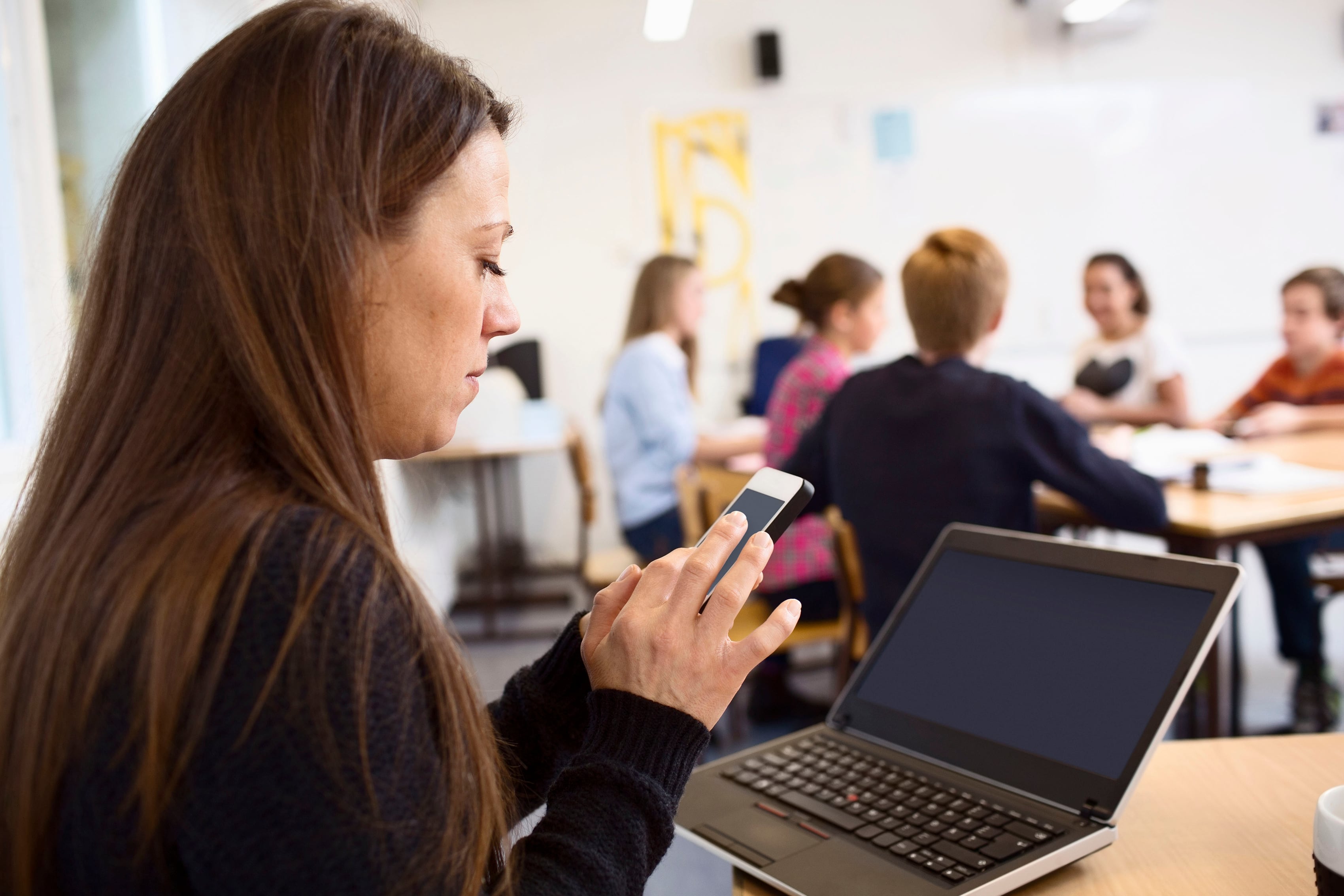 Teacher using mobile phone at classroom with students in background.