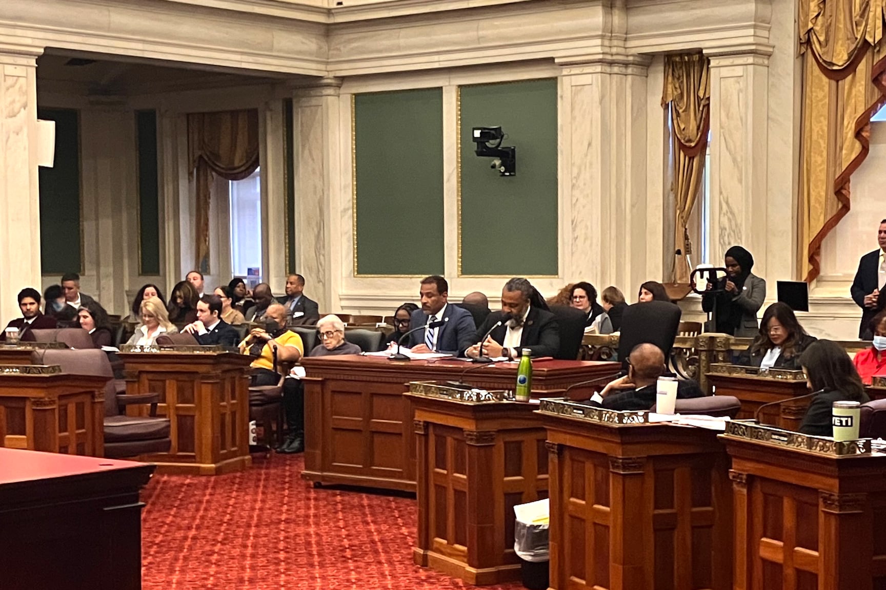 A group of people sit at big wood desks in a room with red carpet.