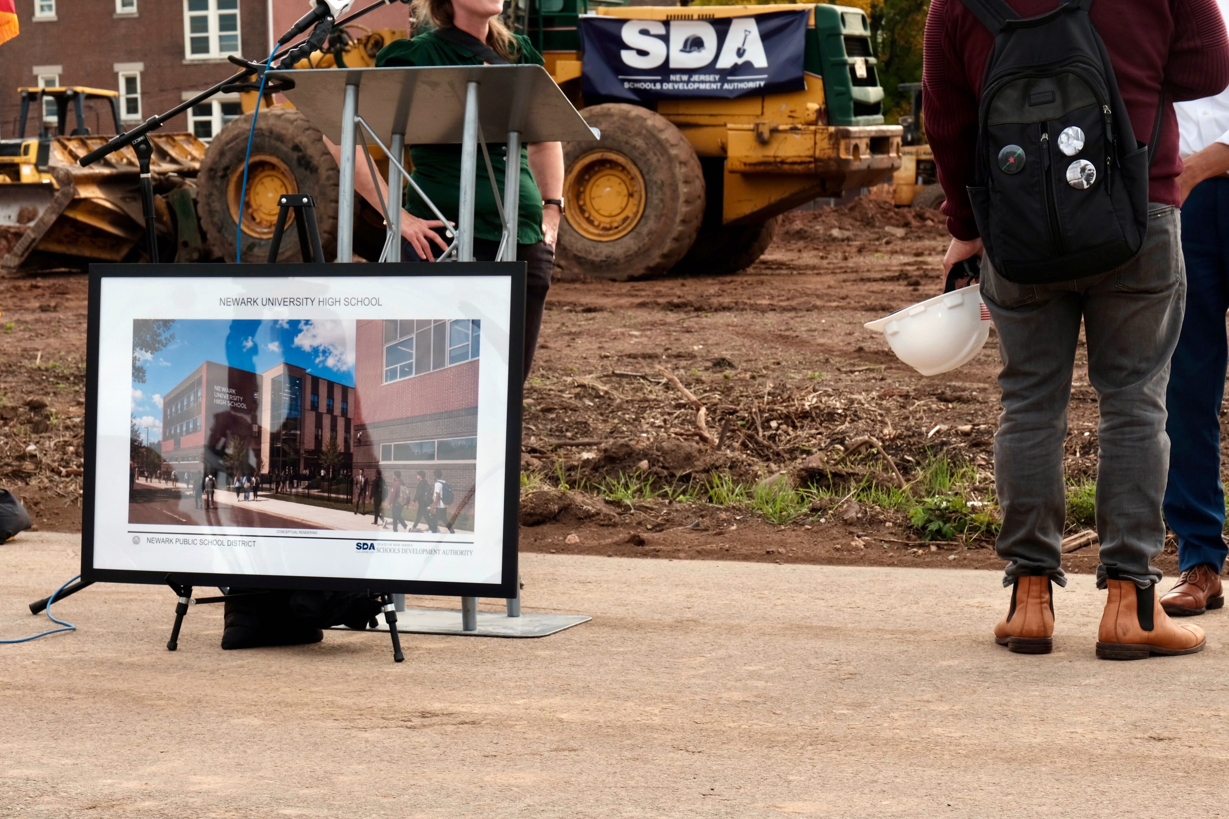 A photograph of a generated model of a school building sits by a construction site with a partial view of a tractor and a person standing nearby.