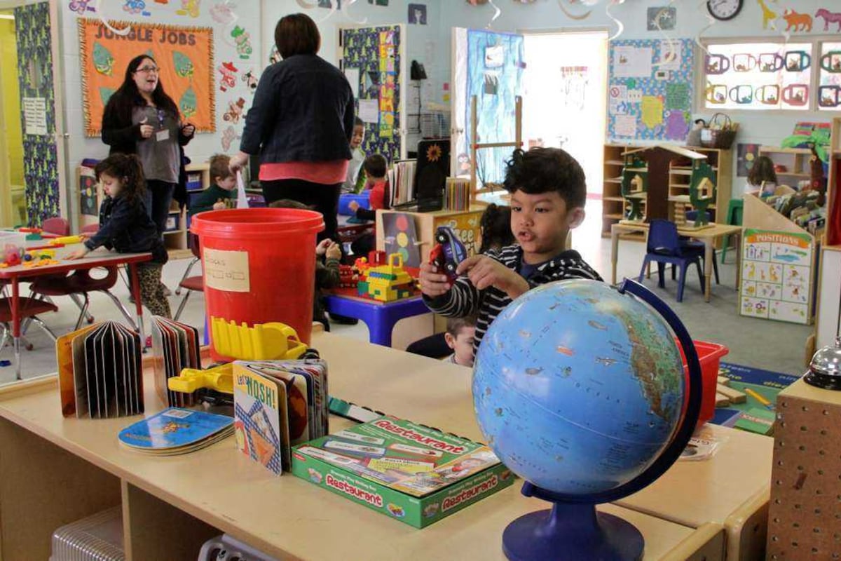 A boy plays in a classroom, just behind a large globe. There are two adults speaking and other kids playing in the classroom.