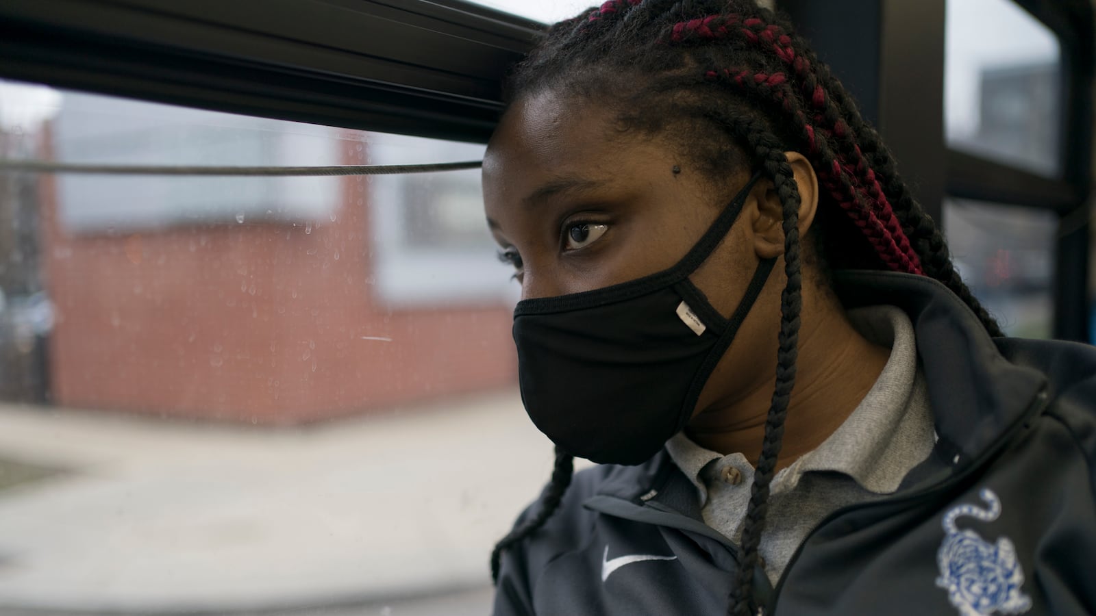 A young woman looks out of a bus window, wearing a black mask with long braided hair. She is wearing a grey track jacket with a school logo near the lapel.