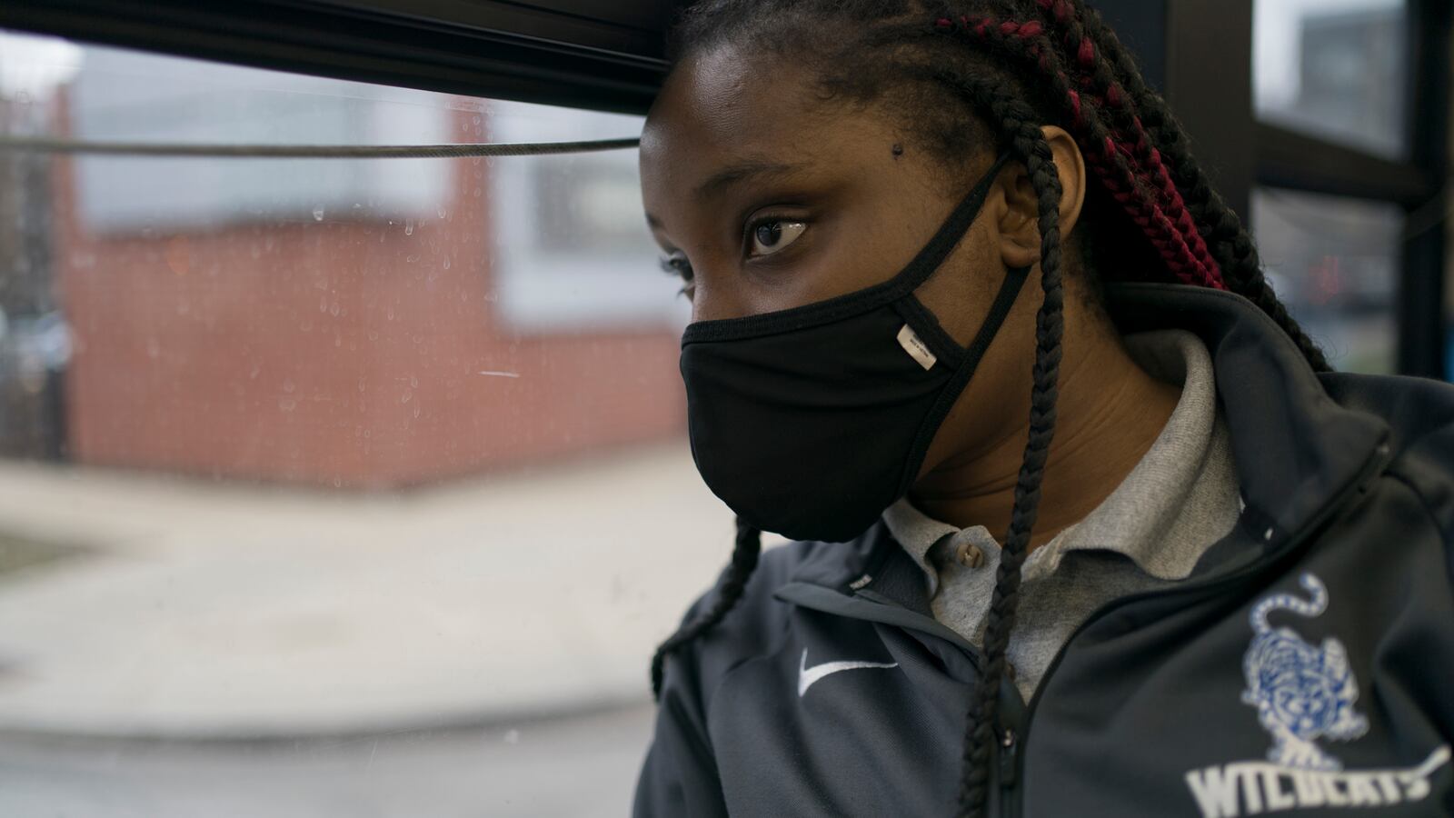 A young woman looks out of a bus window, wearing a black mask with long braided hair. She is wearing a grey track jacket with a school logo near the lapel.