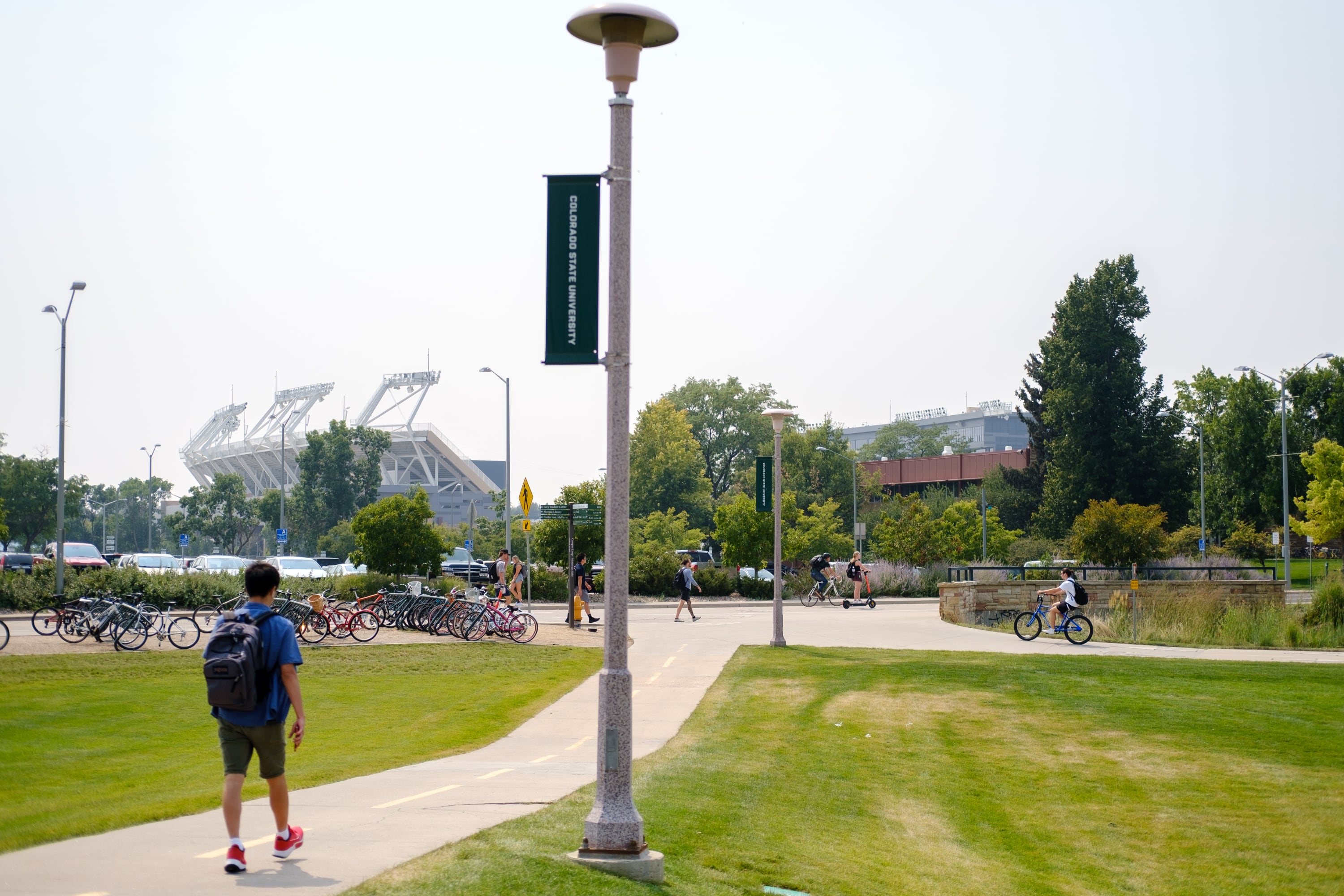 A college student with a black backpack walks on a sidewalk lined with grass and a view of a grey stadium in the background.