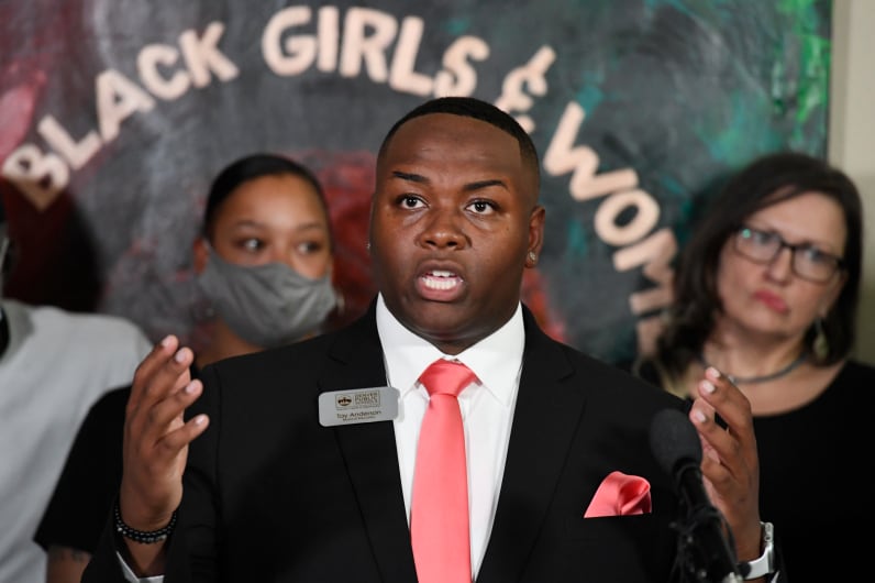 Denver school board member Tay Anderson gestures while speaking at a press conference. He is wearing a black suit and salmon colored tie. Supporters are standing behind him.