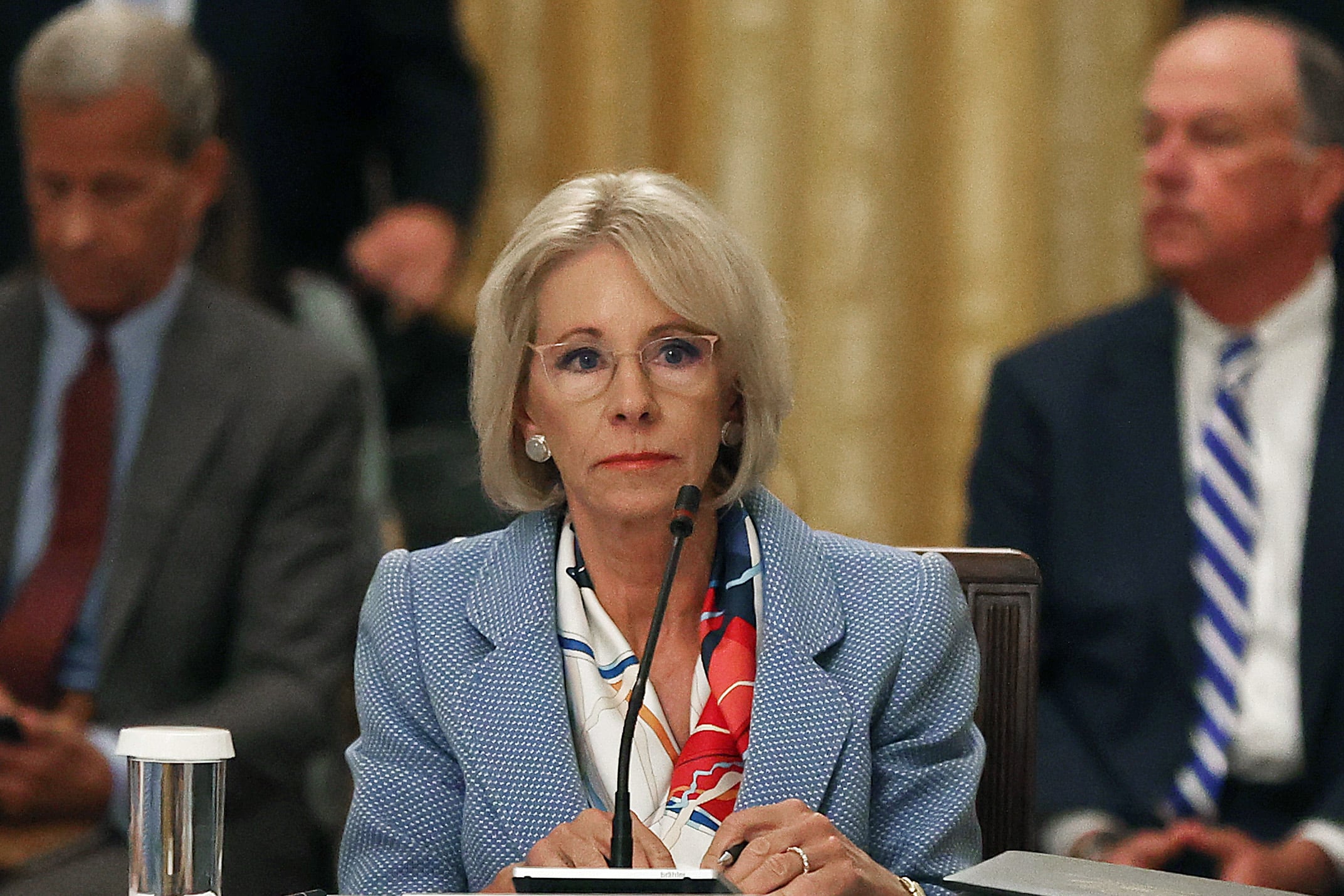 Betsy DeVos wearing a blazer and glasses sits at a table before a microphone