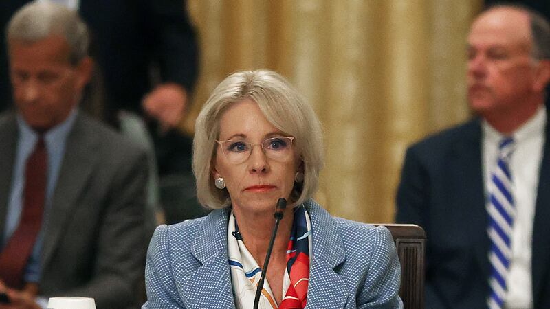Betsy DeVos wearing a blazer and glasses sits at a table before a microphone