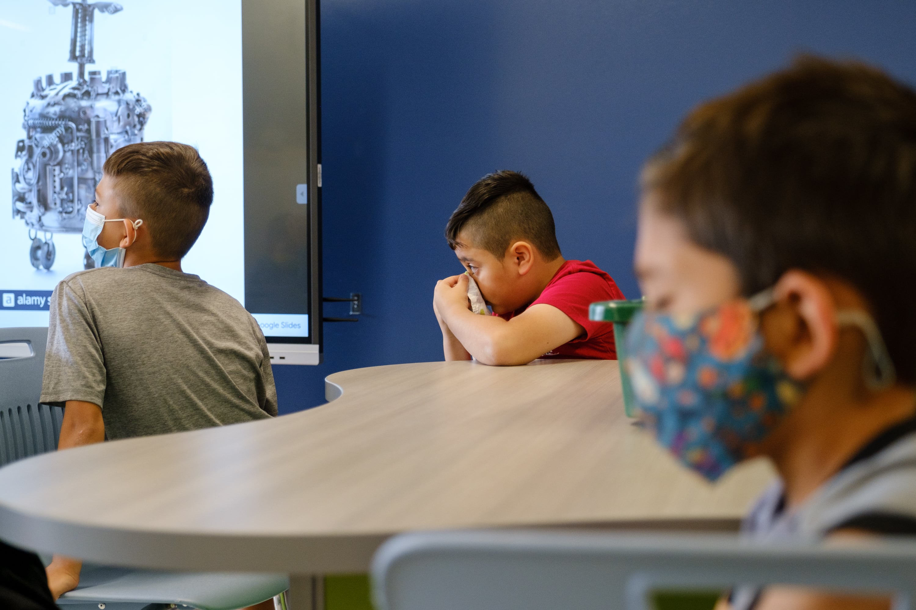 Three elementary-age boys sit at a table in a classroom. All three are wearing masks, though one seems to be holding his in place with his hands.