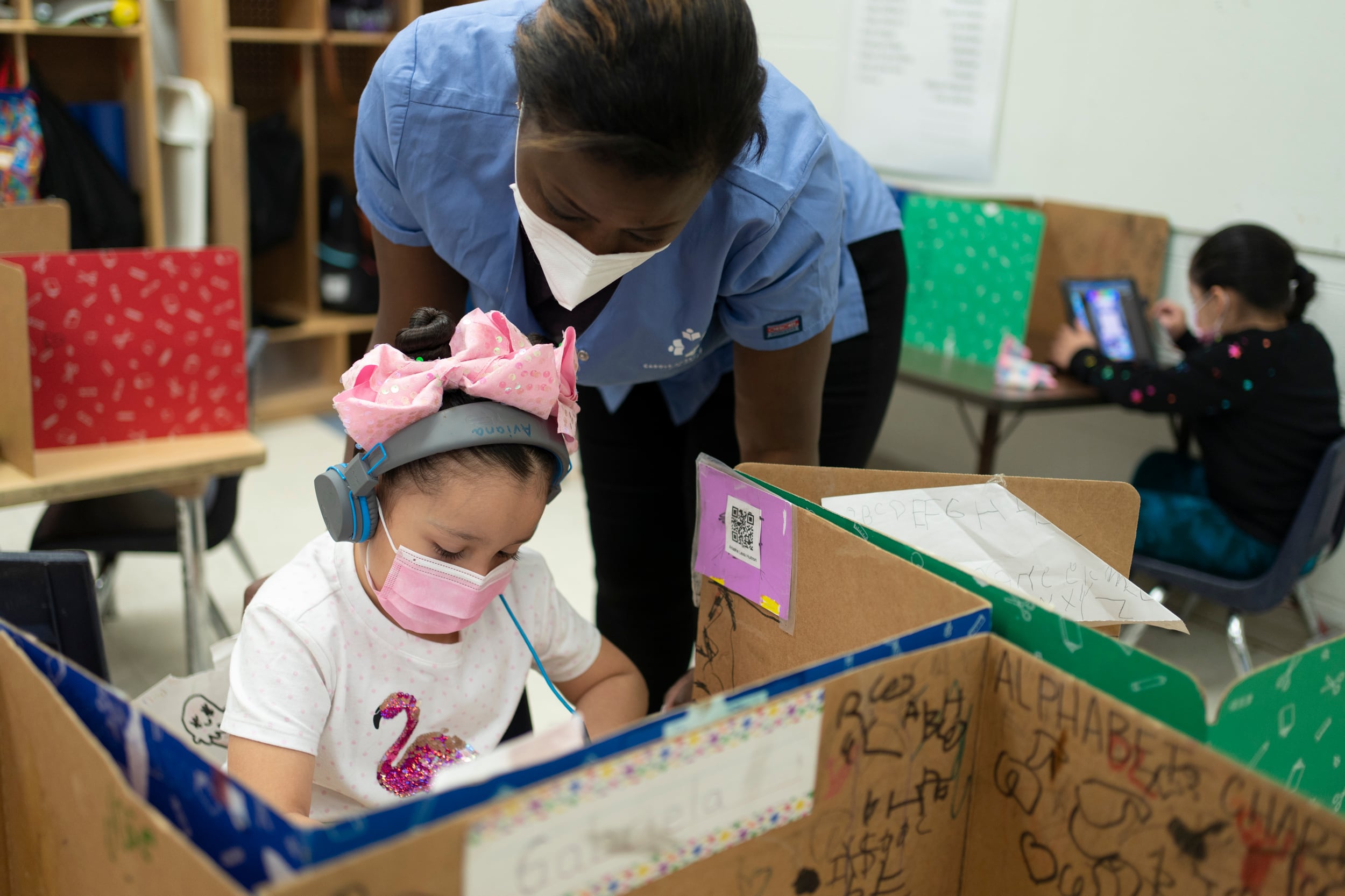 A girl wearing a pink bow, pink protective mask and white shirt with a flamingo works on an assignment with a teacher looking over her shoulder, wearing a blue shirt and KN-95 mask. Another child studies in the background.