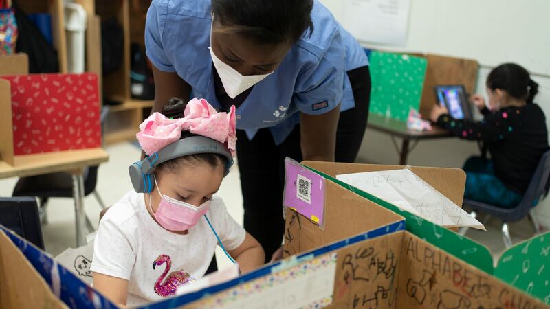 A girl wearing a pink bow, pink protective mask and white shirt with a flamingo works on an assignment with a teacher looking over her shoulder, wearing a blue shirt and KN-95 mask. Another child studies in the background.
