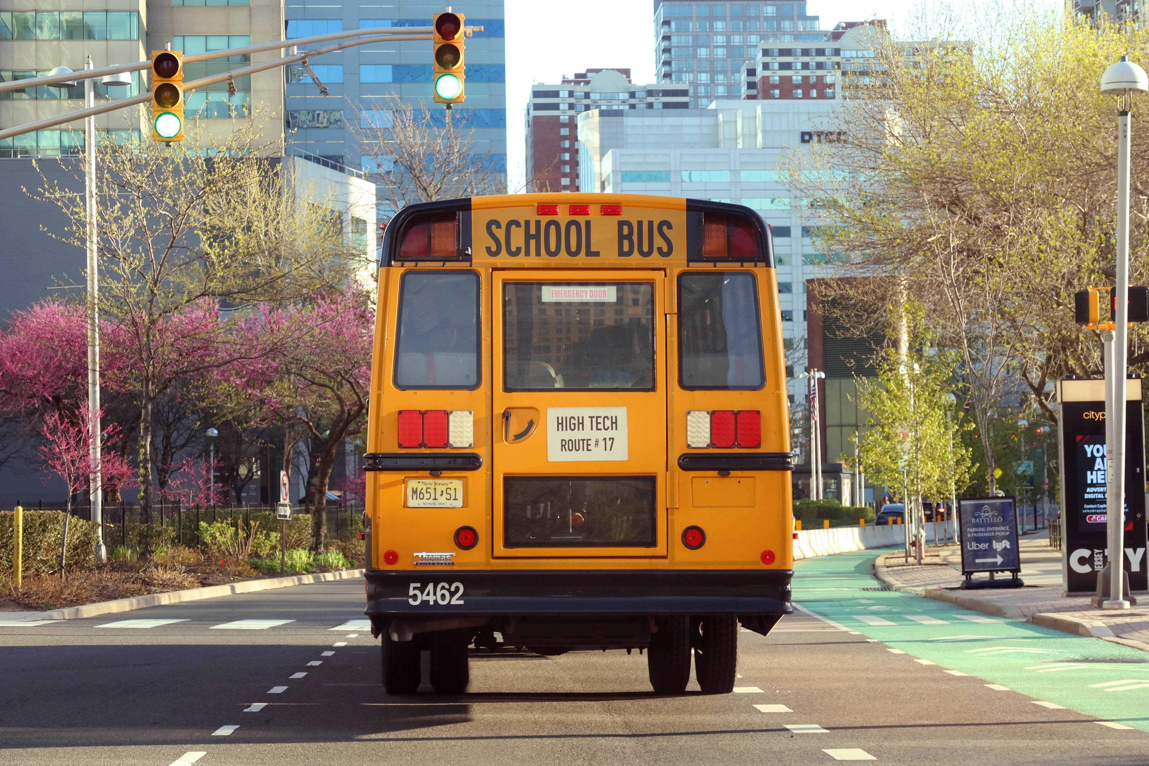 A yellow and black school bus drives away from the camera down a street with a cityscape in the background.