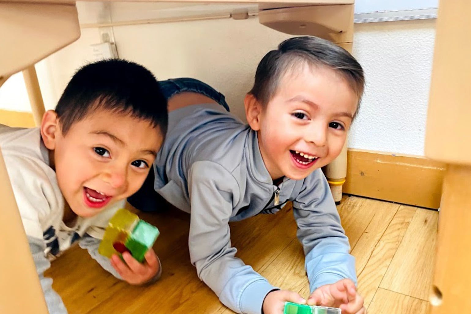 Two young boys smile while crouched under a table on a wood floor.