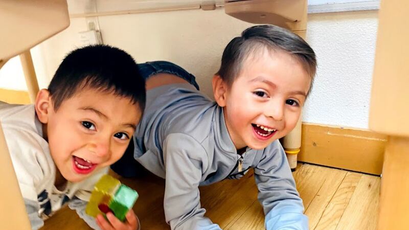 Two young boys smile while crouched under a table on a wood floor.