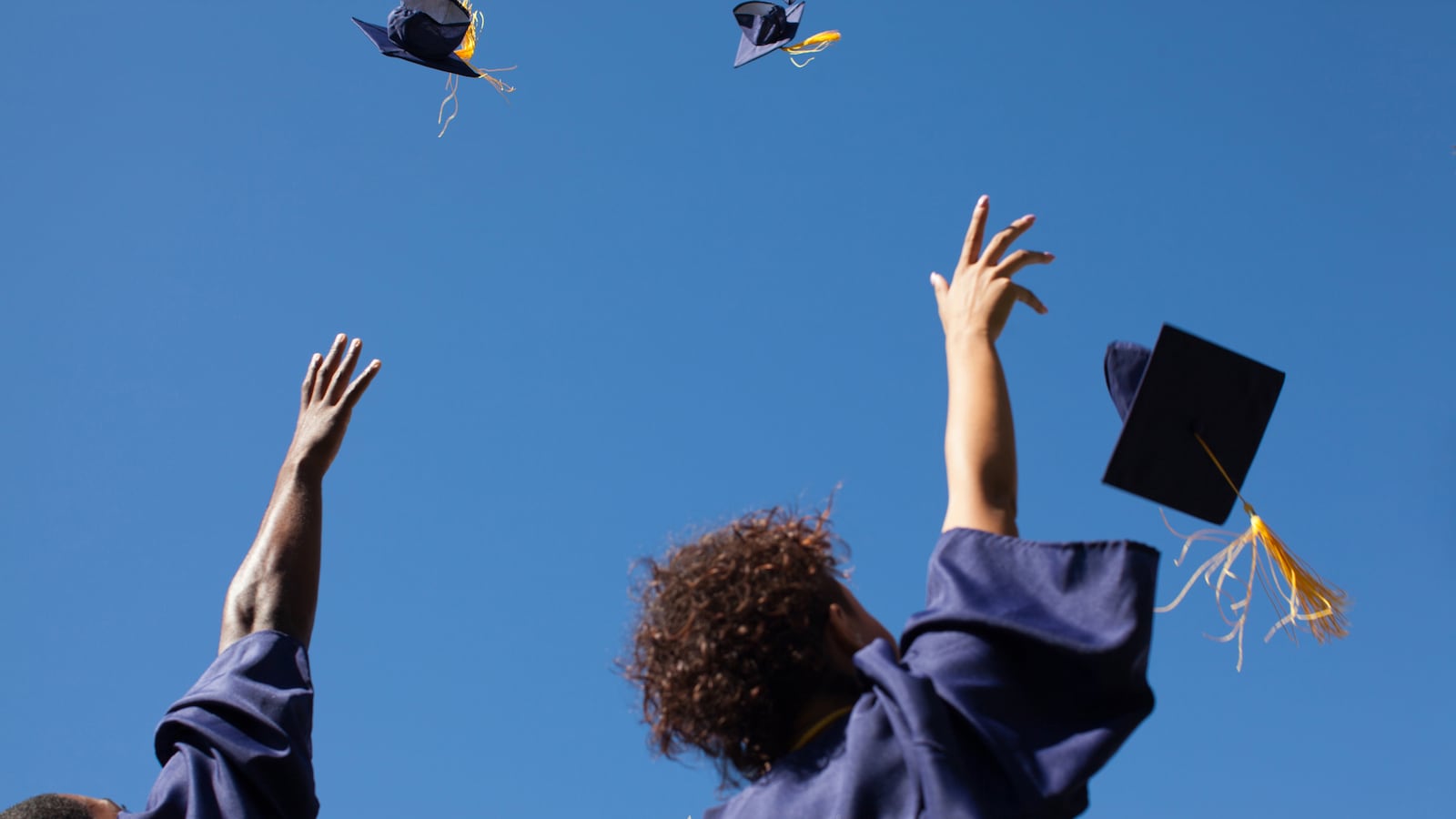 Two high school graduates wearing blue gowns throw their caps into the air.