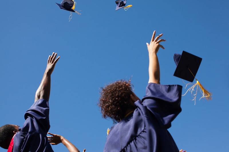 Two high school graduates wearing blue gowns throw their caps into the air.