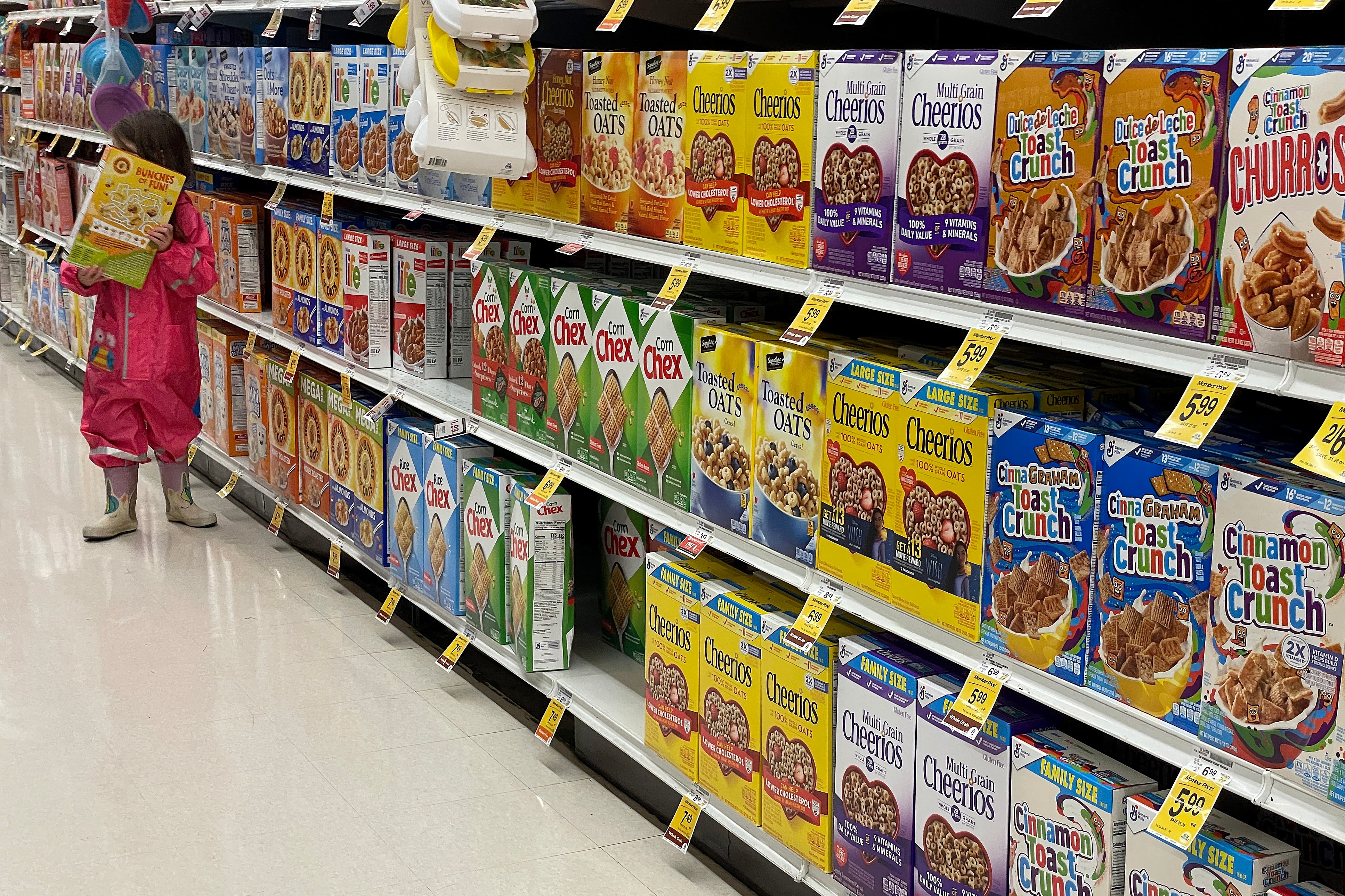 A young child carries a large box of cereal covering their face while they stand next to an aisle full of cereal boxes.