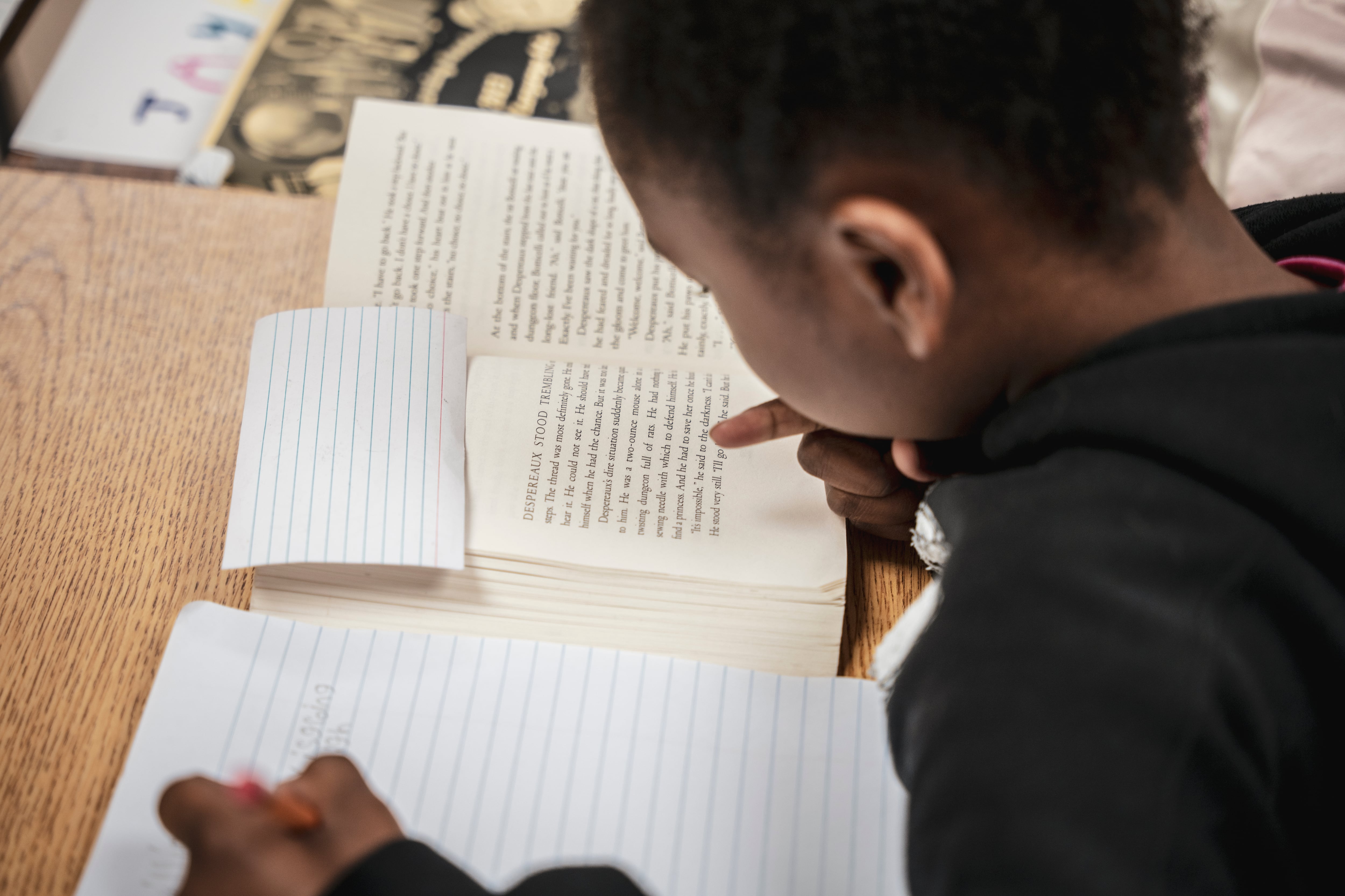 A photograph of a Black student looking down and writing in a notebook at a wooden school desk.