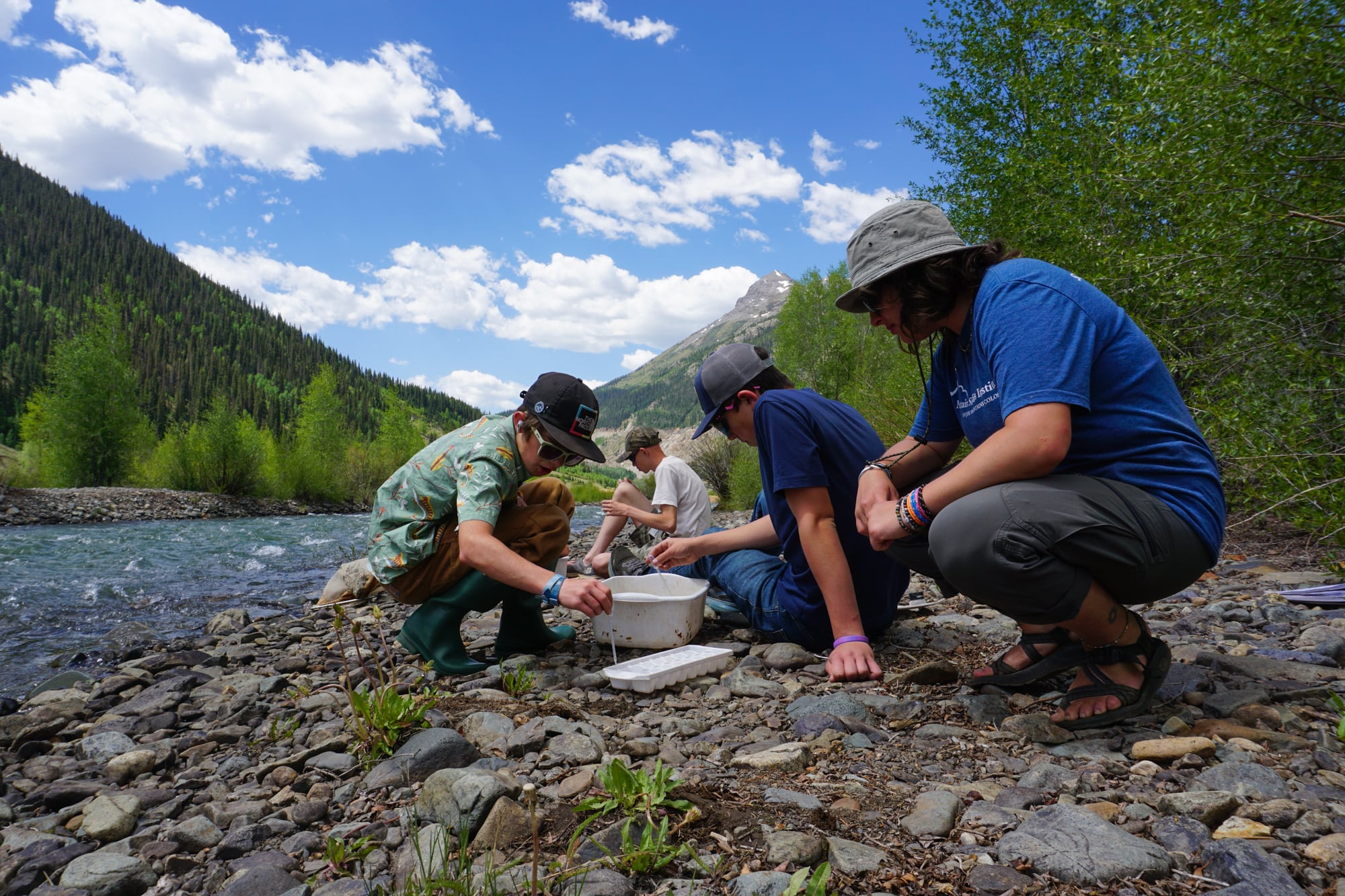 Young teenagers and a teacher in outdoor gear crouch in the rocks next to a stream. They are looking through the rocks, and they have a plastic bucket.