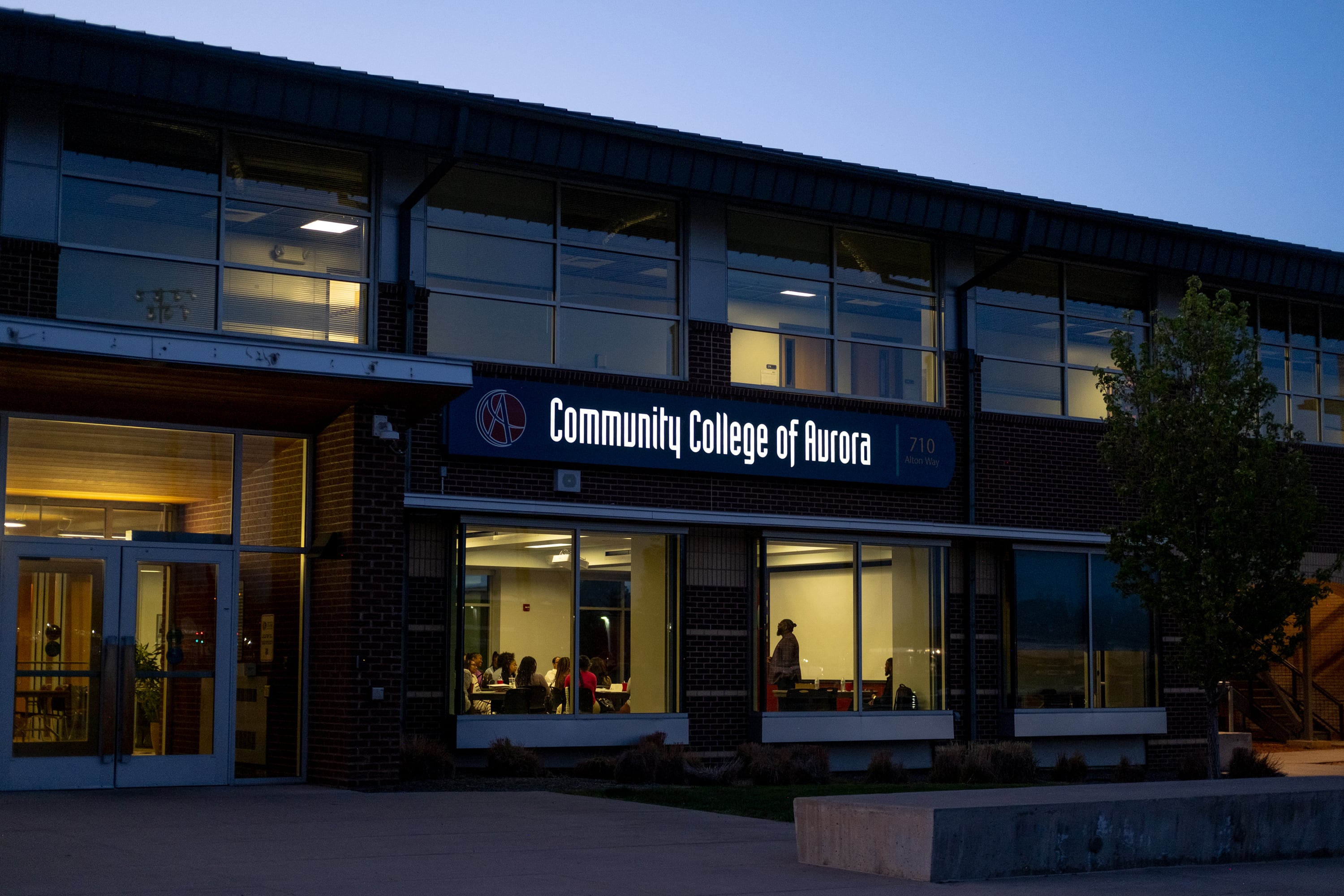 A few people stand inside of a building that is illuminated while the sunsets.