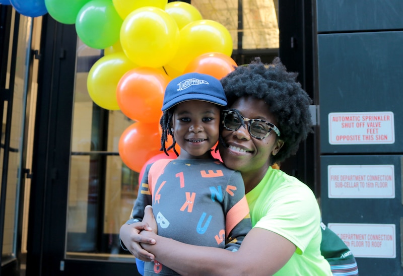 A mom with short dark hair poses for a photograph with her young son in front of a balloon arch outside.