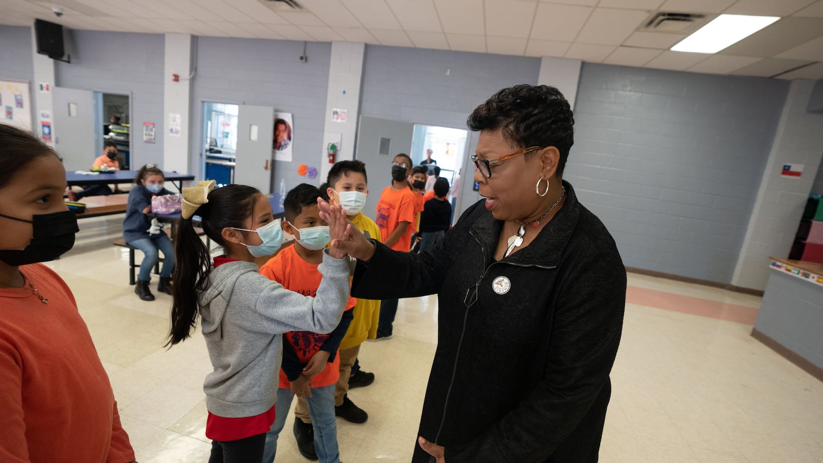A woman wearing glasses gives high fives to a line of students in a school cafeteria