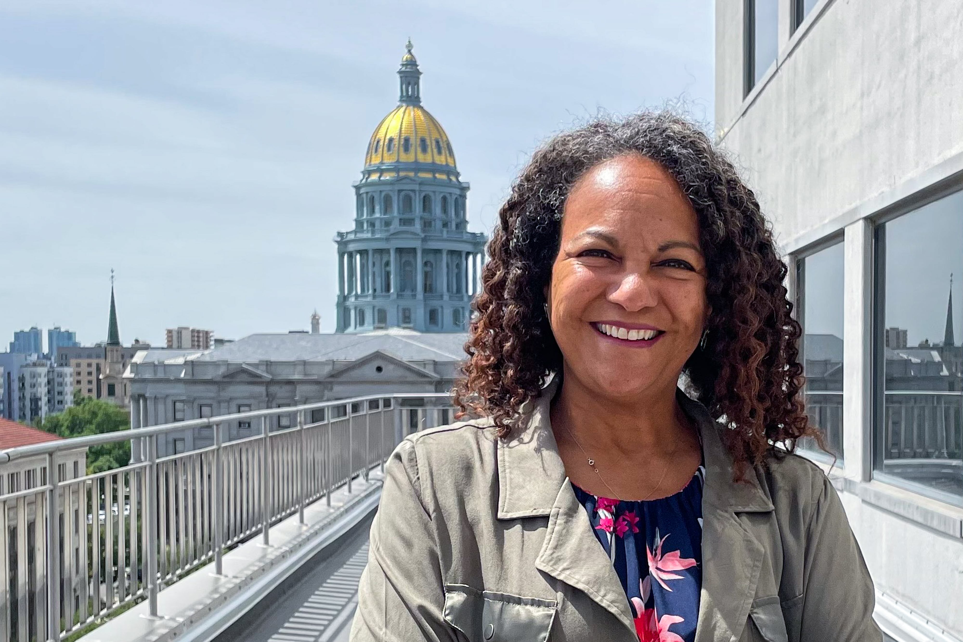 A woman with curly brown hair smiles for a portrait, standing on a balcony with the Colorado State Capitol behind her.