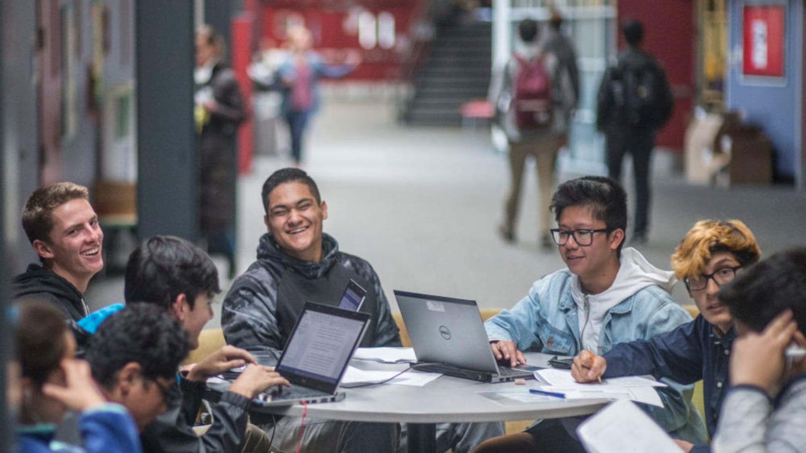 A group of students in hoodies work around a table in a hallway or common area of DSST: Montview in 2017. They appear to be mostly boys and they’re smiling and laughing. Several laptops are open on the table.