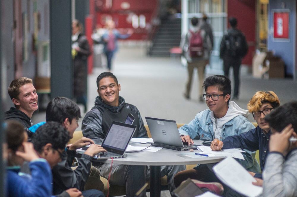 A group of students in hoodies work around a table in a hallway or common area of DSST: Montview in 2017. They appear to be mostly boys and they’re smiling and laughing. Several laptops are open on the table.