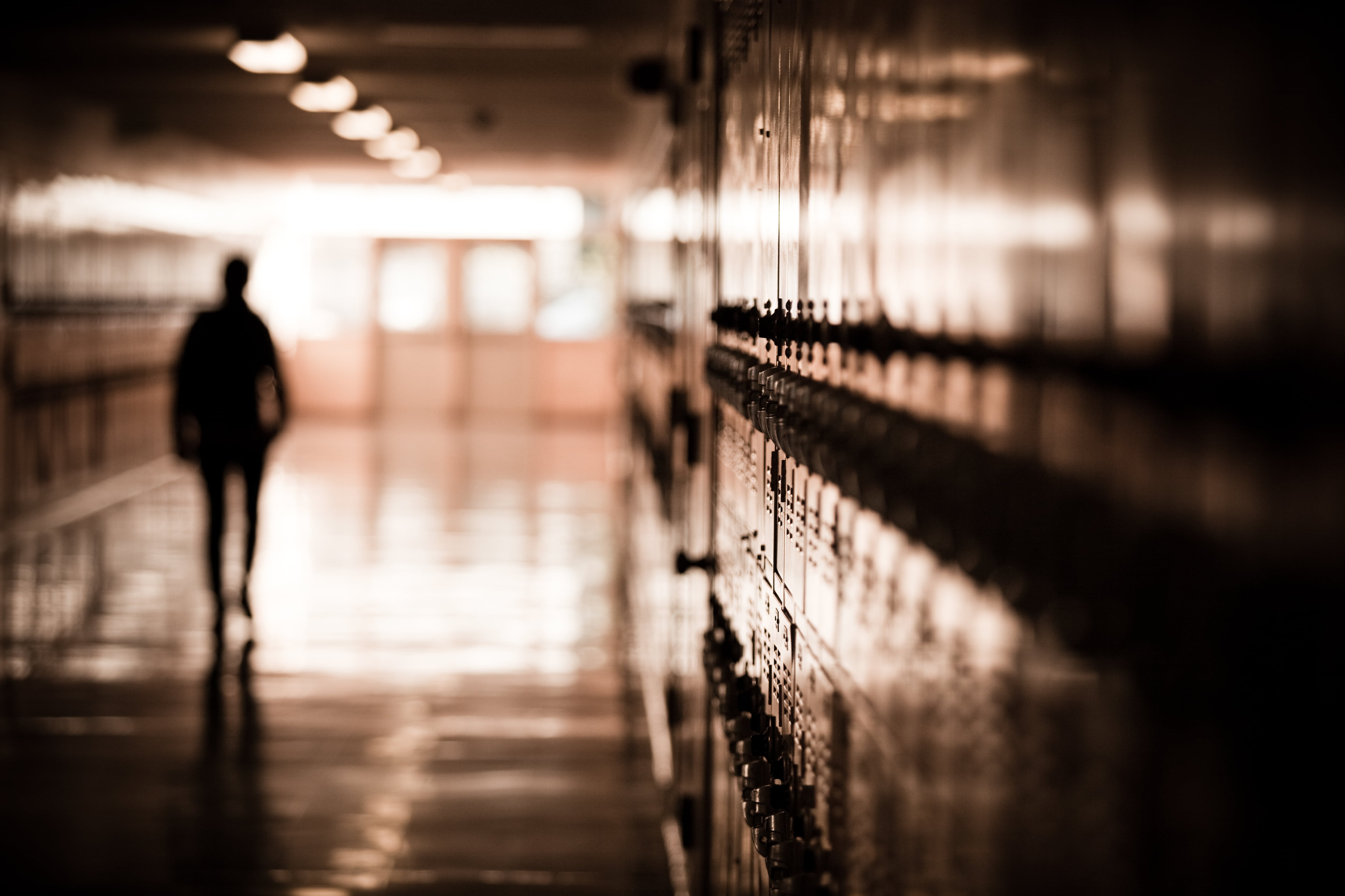 A high school student walks down a dark hallway in a public high school, silhouetted by daylight spilling in and reflecting off of the floor and lockers.