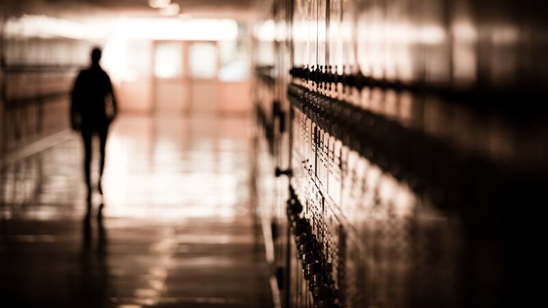 A high school student walks down a dark hallway in a public high school, silhouetted by daylight spilling in and reflecting off of the floor and lockers.