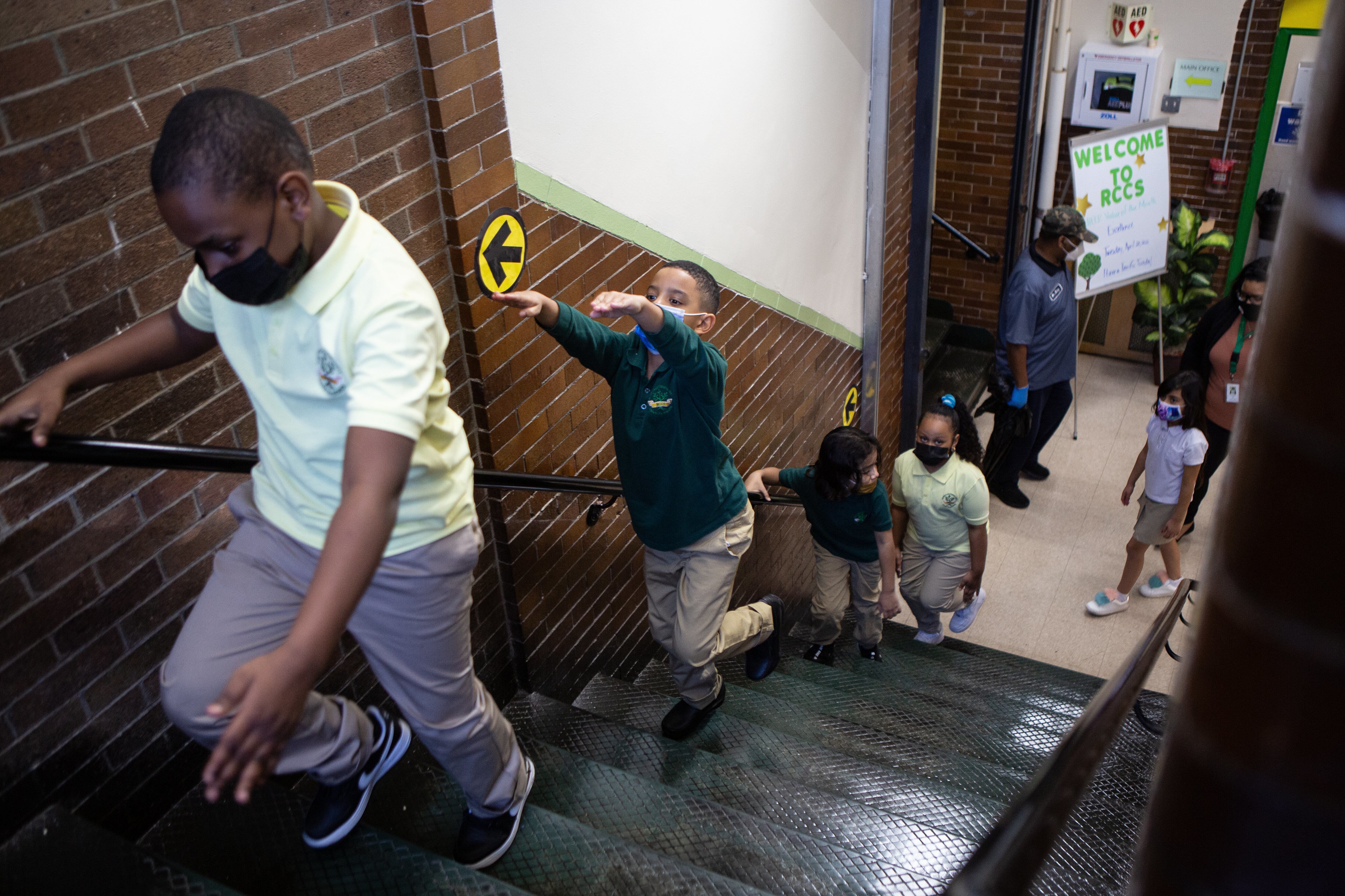 Masked second grade students walk up a stairwell, with one boy raising his arms in the air to practice arm’s-length social distancing.