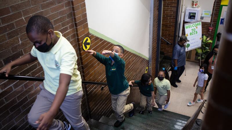 Masked second grade students walk up a stairwell, with one boy raising his arms in the air to practice arm’s-length social distancing.