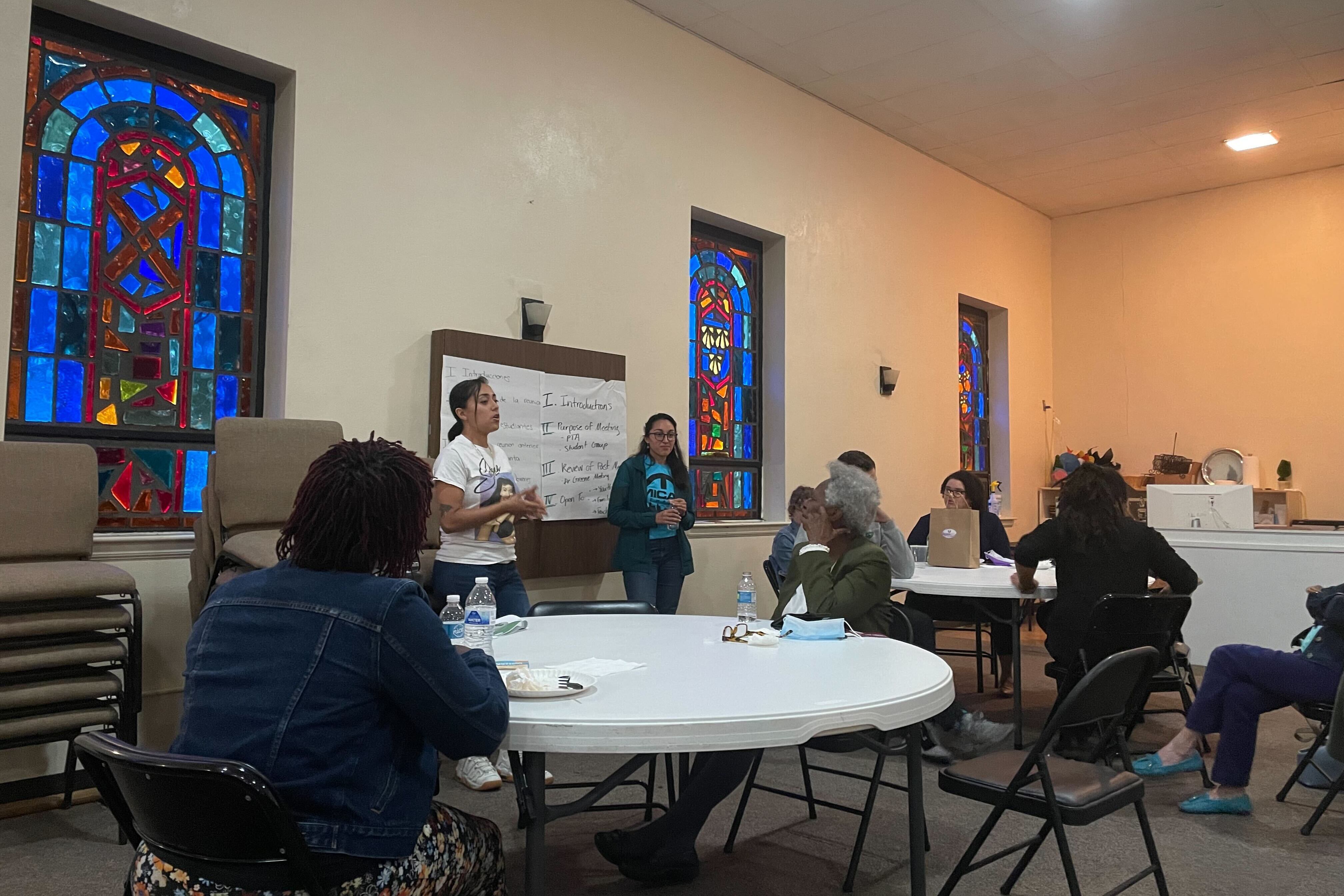 Two women stand in front of a crowd of people sitting at tables in an old church building with stained glass windows