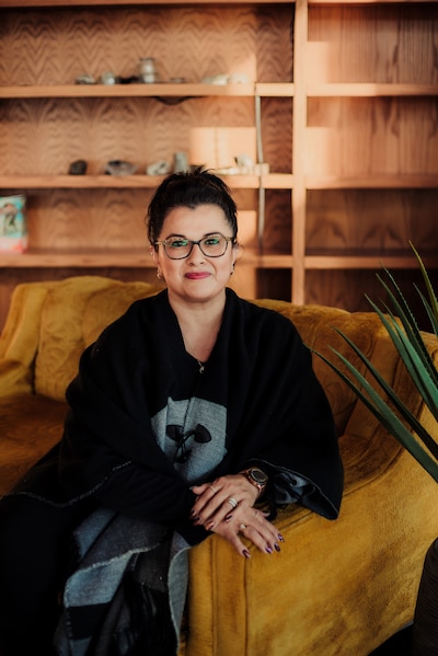 A woman with dark hair tied up poses for a portrait on a sofa and in font of a book case.