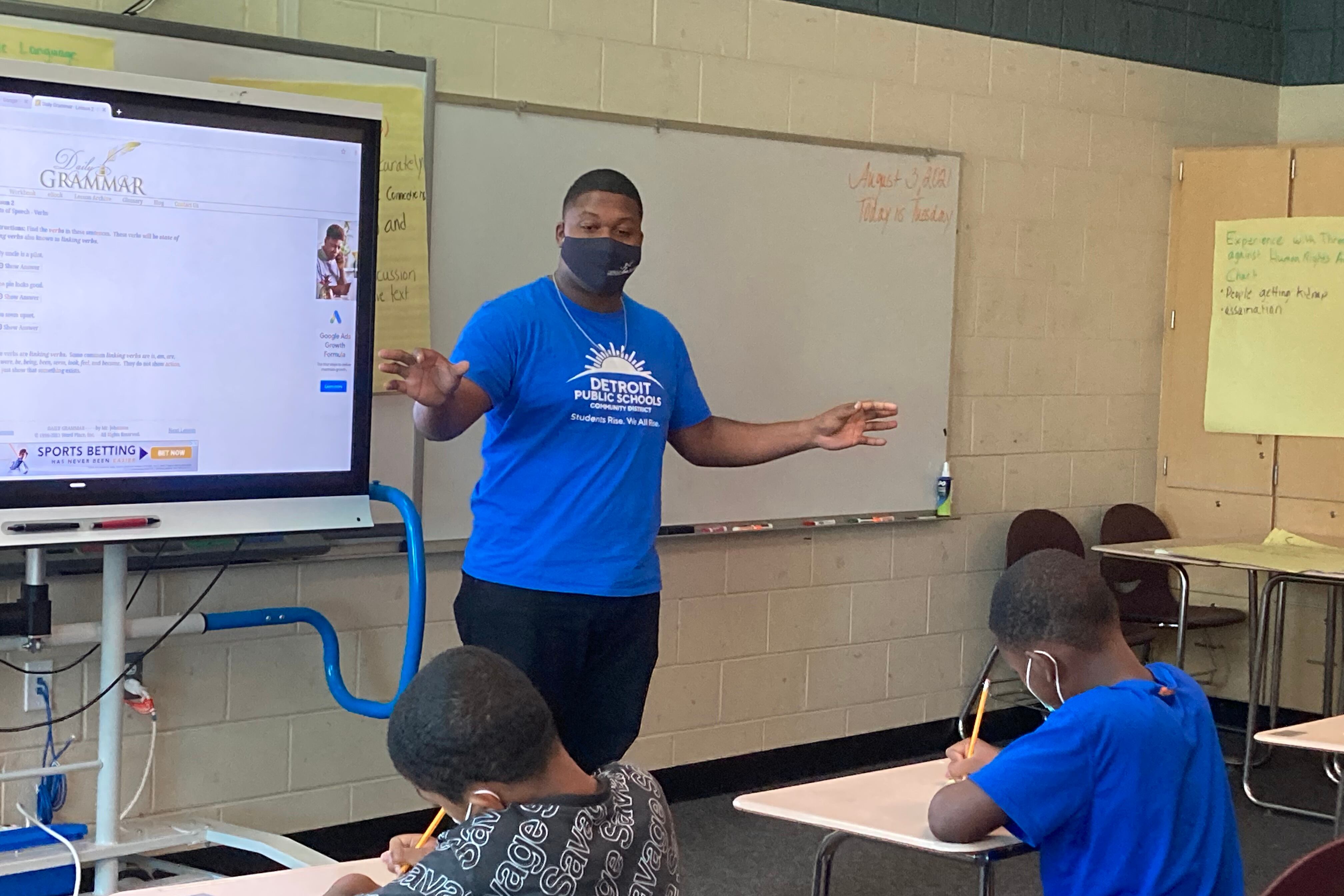 Man in blue t-shirt stands at front of classroom, next to a projection screen, instructing students.