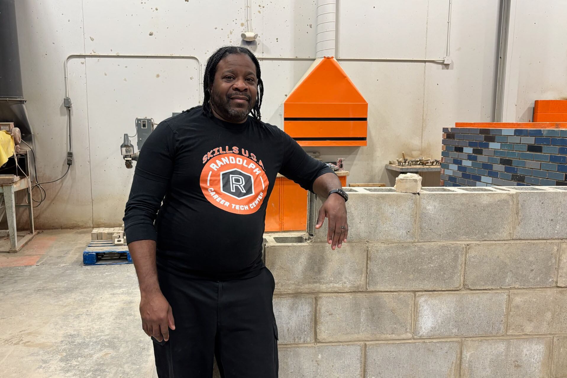 A Black man in a black t-shirt stands near a concrete brick wall in a warehouse.