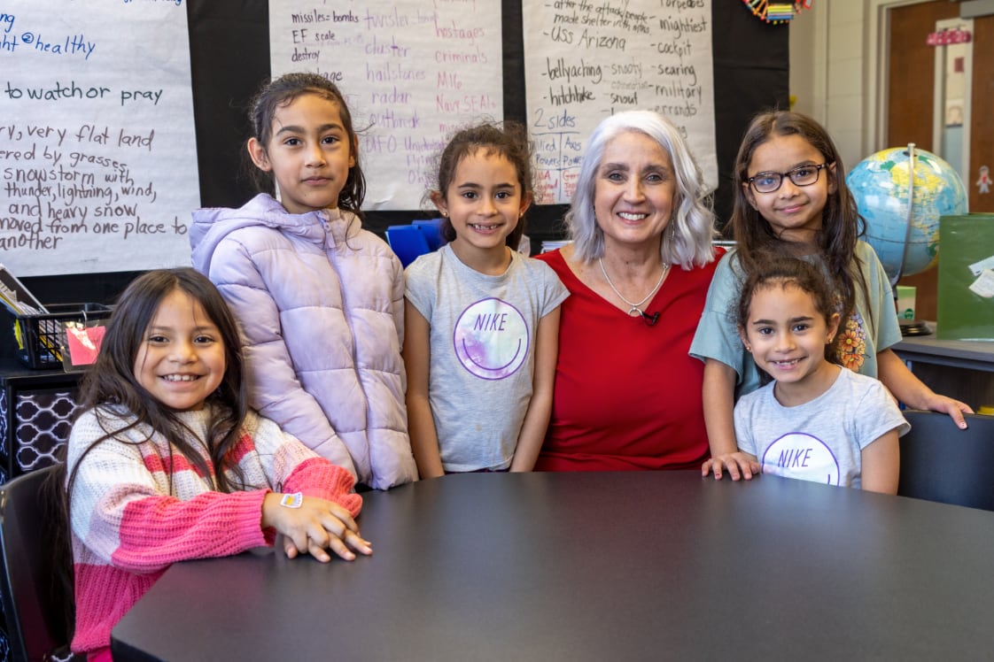 Five young students stand next to a an adult teacher wearing a red shirt in a classroom.